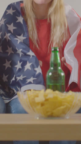 Vertical-Video-Of-Woman-Wrapped-In-American-Flag-Celebrating-Watching-Sports-Game-On-TV-At-Home-